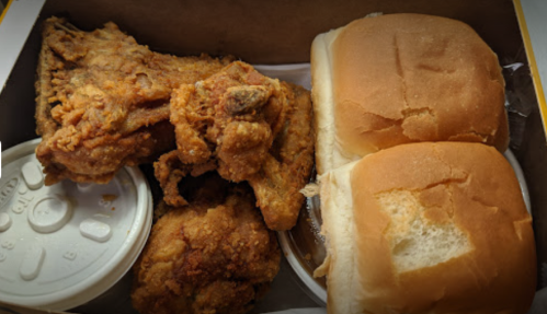 A takeout box containing fried chicken, two soft buns, and a cup of dipping sauce.