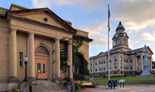 Historic buildings with classical architecture, featuring a clock tower and a statue, set against a cloudy sky.