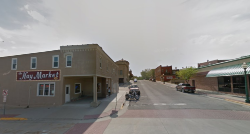 A street view of a small town featuring shops, parked cars, and a clear sky.