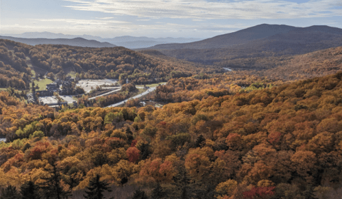 A scenic view of rolling hills covered in autumn foliage, with a winding road and distant mountains under a cloudy sky.