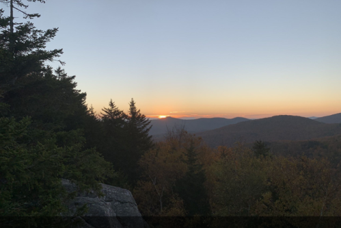 A serene mountain landscape at sunset, with trees in the foreground and rolling hills in the background.