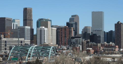 A skyline view of Denver, Colorado, featuring modern skyscrapers and a green bridge in the foreground.