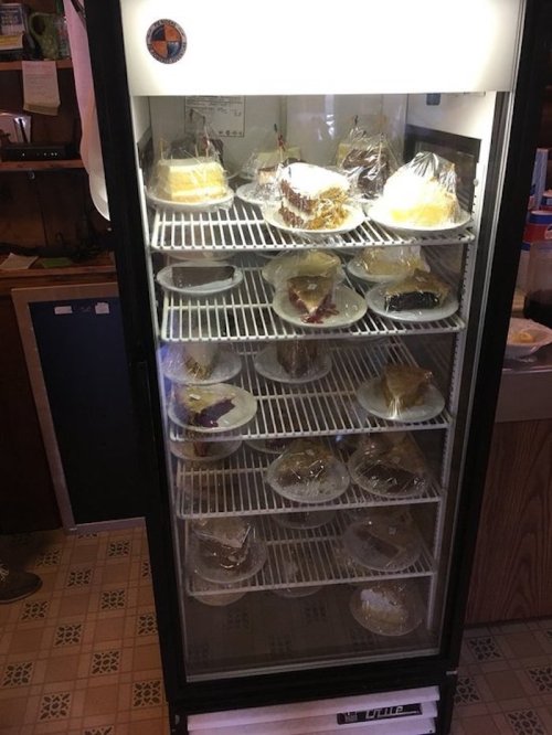 A glass-front refrigerator displaying various cakes and desserts on plates, all covered with plastic wrap.