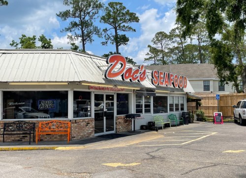 Exterior of Doc's Seafood restaurant with a sign, outdoor seating, and trees in the background on a sunny day.