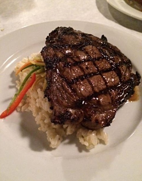 A grilled steak with a charred surface served on a bed of rice, garnished with colorful vegetables.
