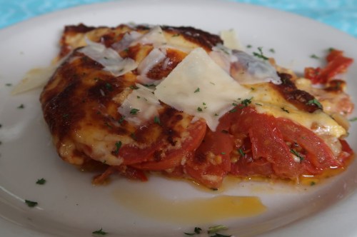 A slice of baked dish with layers of tomatoes, cheese, and herbs on a white plate.