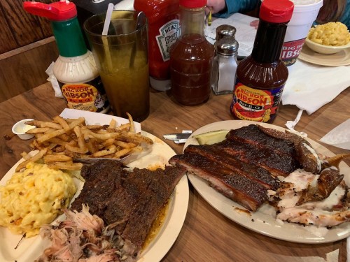 A plate of barbecue ribs, pulled pork, and mac and cheese, with fries and various condiments on a wooden table.