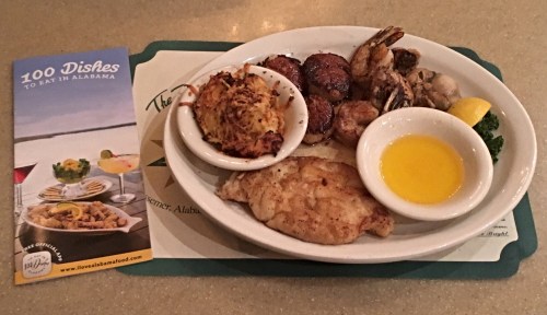 A seafood platter featuring fried fish, shrimp, crab cakes, and a lemon wedge, alongside a brochure about Alabama dishes.