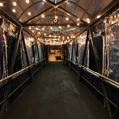 A dimly lit walkway with string lights, lined with transparent curtains, leading to an entrance.