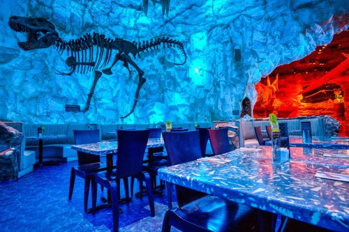 A dimly lit restaurant with blue lighting, featuring a dinosaur skeleton on the wall and marble tables.