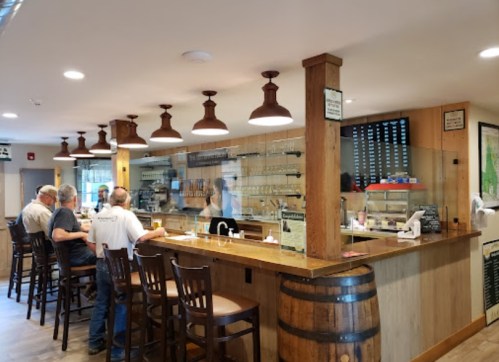 A cozy bar with wooden stools, patrons seated, and a glass display showcasing drinks and snacks. Warm lighting enhances the atmosphere.