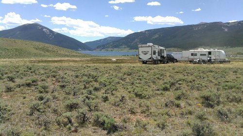 A scenic view of two RVs parked near a lake, surrounded by mountains and grassy terrain under a blue sky.
