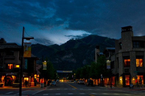 A quiet street at dusk, lined with shops and trees, with mountains silhouetted against a cloudy sky.
