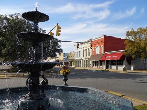 A decorative fountain in the foreground with a street and historic buildings in the background under a blue sky.