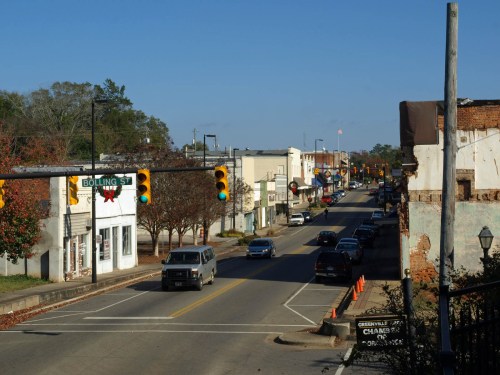 A view of a small town street with shops, traffic lights, and trees, under a clear blue sky.