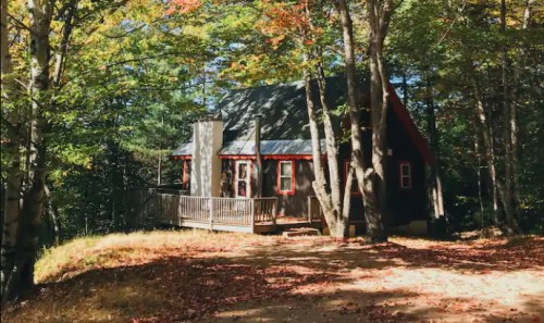 A cozy cabin surrounded by colorful autumn trees, with a wooden deck and a gravel path leading up to it.