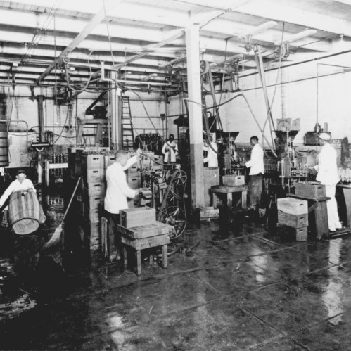 Black and white photo of workers in a factory, operating machinery and handling materials in a large industrial space.
