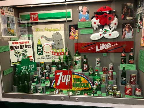 Display case featuring vintage 7UP and Like Cola memorabilia, including bottles, signs, and a plush mascot.
