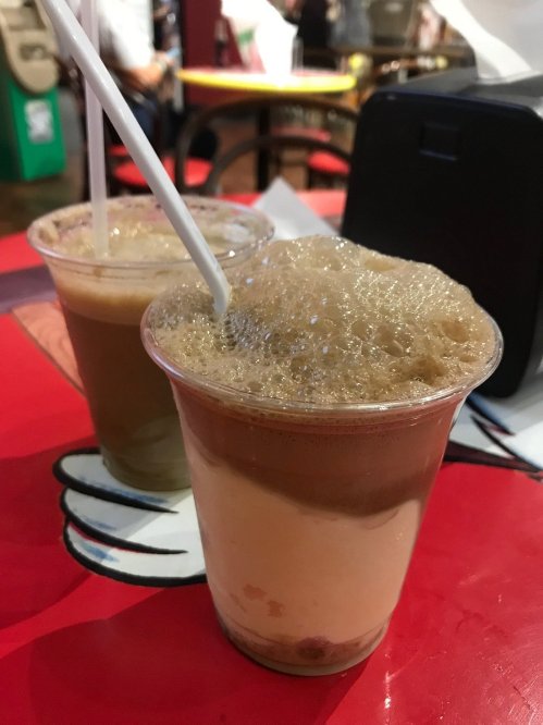 Two cups of frothy beverages on a red table, one light brown and one darker, with straws inserted.