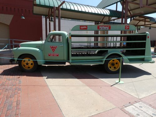 A vintage green truck with an open bed, displaying retro signage, parked in a public area with brick pavement.