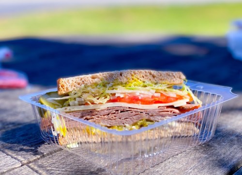 A close-up of a sandwich with layers of lettuce, tomato, and sliced meat, served in a clear plastic container.