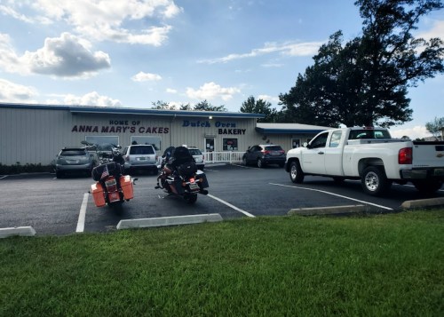A bakery building with a sign reading "Home of Anna Mary's Cakes," surrounded by parked cars and motorcycles.