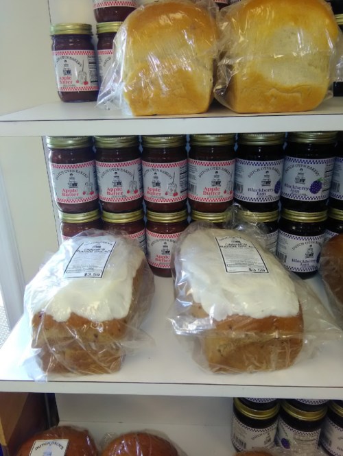 Two loaves of bread wrapped in plastic on a shelf, with jars of preserves in the background.