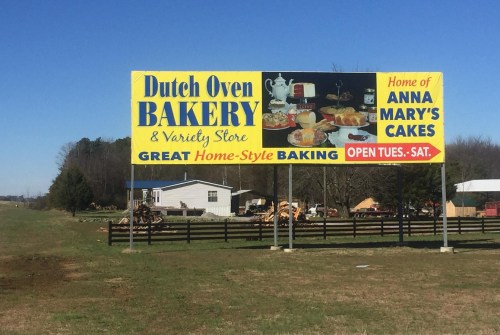 A colorful billboard for Dutch Oven Bakery and Variety Store, featuring baked goods and promoting Anna Mary's Cakes.