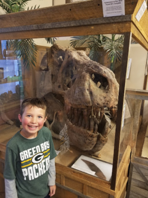 A young boy smiles next to a large dinosaur skull displayed in a glass case.
