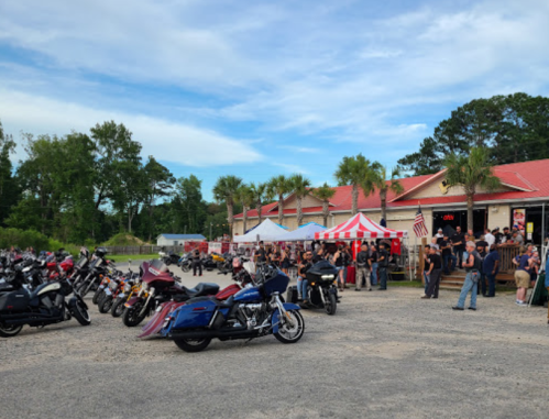 A lively motorcycle gathering outside a venue, with numerous bikes parked and people socializing under a blue sky.