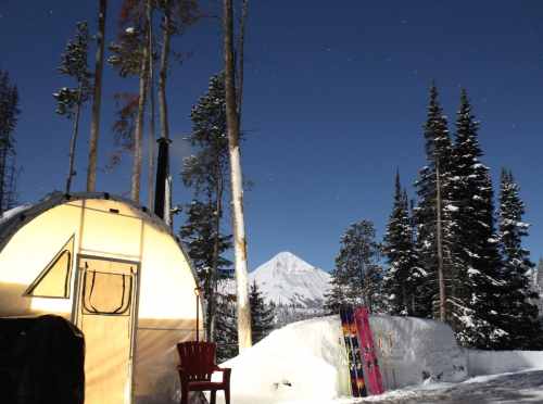 A cozy tent in a snowy landscape, surrounded by tall trees and mountains under a starry sky, with skis nearby.