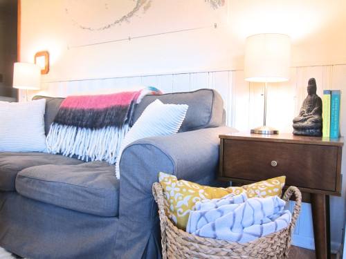 Cozy living room scene with a gray sofa, colorful throw, side table with a lamp, and a basket of blankets.