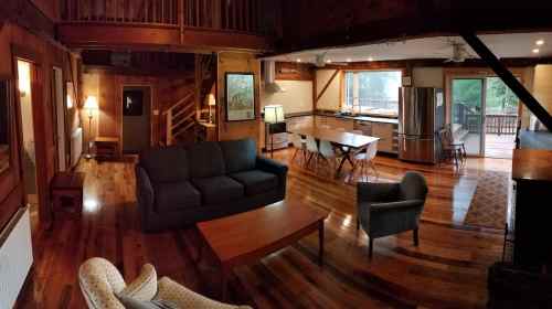 Cozy wooden interior of a spacious living area with a sofa, chairs, and a kitchen, featuring large windows and natural light.