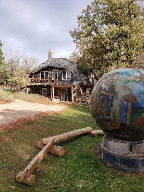 A rustic wooden house with a unique roof, surrounded by trees, and a large spherical sculpture in the foreground.