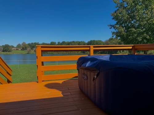 A wooden deck with a hot tub overlooking a serene lake and clear blue sky.