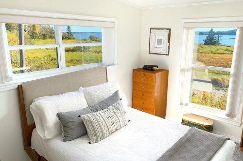 A cozy bedroom with a bed, decorative pillows, and large windows showcasing a scenic view of nature outside.