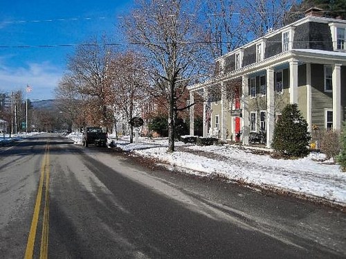 A snowy street scene featuring a large house with a red door, trees, and a clear blue sky.