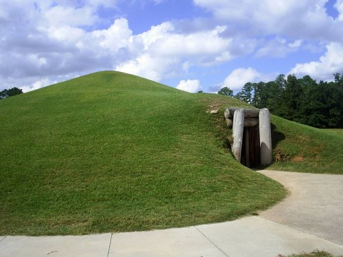 A grassy mound with a wooden entrance, surrounded by trees and a cloudy sky.