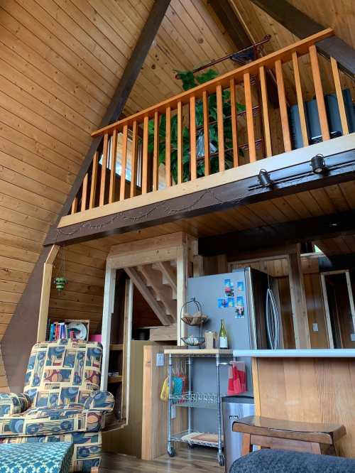 Cozy A-frame cabin interior with wooden walls, a loft, and a living area featuring a patterned chair and kitchen appliances.