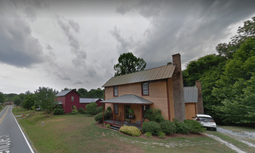 A two-story wooden house with a metal roof, surrounded by greenery, near a road and a red barn in the background.