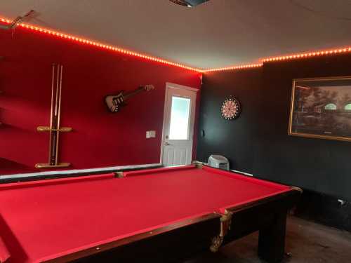 A game room featuring a red pool table, dartboard, guitar decor, and ambient lighting on the walls.