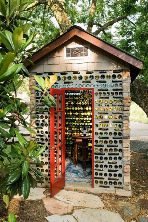 A small, charming building made of glass bottles, featuring a red door and a cozy interior with a table and chairs.