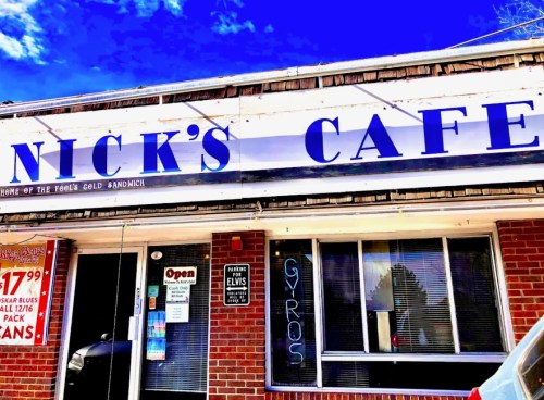 A storefront of Nick's Cafe with a blue sign, open window, and a neon "Gyros" sign. Bright blue sky above.