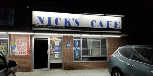 A nighttime view of Nick's Cafe, featuring a sign and a parked car outside a brick building.