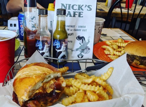 A plate with a burger and crinkle-cut fries, alongside hot sauce bottles and a menu at Nick's Cafe.