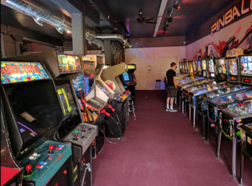 A vibrant arcade room filled with classic pinball machines and arcade cabinets, with a person playing in the background.