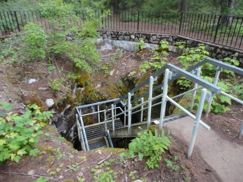 A metal staircase leads down into a rocky, moss-covered pit surrounded by greenery and a railing.