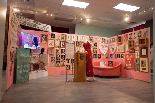 A colorful exhibit featuring framed photos, a red dress on a mannequin, and a pink couch in a museum setting.