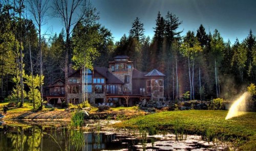 A scenic view of a large house by a pond, surrounded by lush trees and sunlight filtering through the forest.