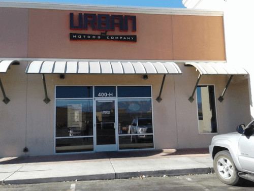 Exterior of Urban Hotdog Company with a large sign, glass doors, and awnings on a sunny day.